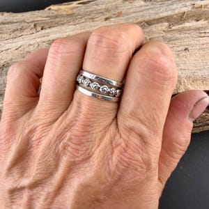 May include: A silver ring set on a hand. The ring features three bands, with the middle band adorned with small, round, textured embellishments. The bands have a polished finish, reflecting light. The ring is worn on a finger, showcasing its design.