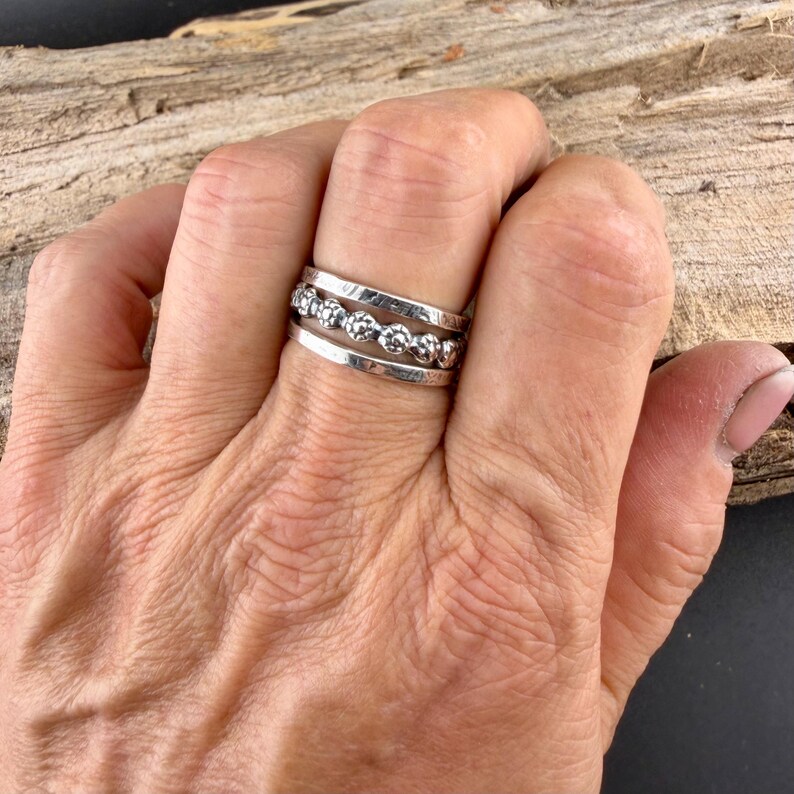 May include: A silver ring set on a hand. The ring features three bands, with the middle band adorned with small, round, textured embellishments. The bands have a polished finish, reflecting light. The ring is worn on a finger, showcasing its design.