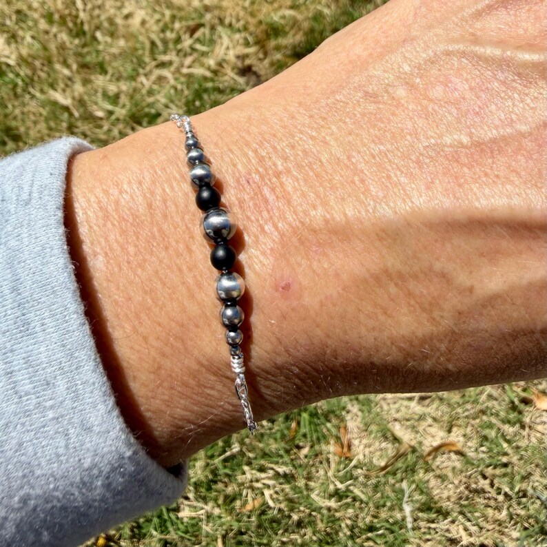 May include: A silver bracelet with a series of beads in varying sizes. The beads alternate between shiny silver and matte black. The bracelet is on a person's wrist, with a light blue sleeve visible in the background.