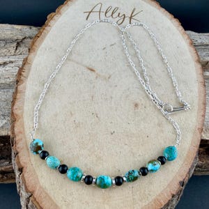 May include: A silver chain necklace with turquoise and black beads. The necklace features a series of round turquoise beads interspersed with smaller black beads. The chain is secured with a simple clasp. The name "Allyk" is written in cursive at the top.