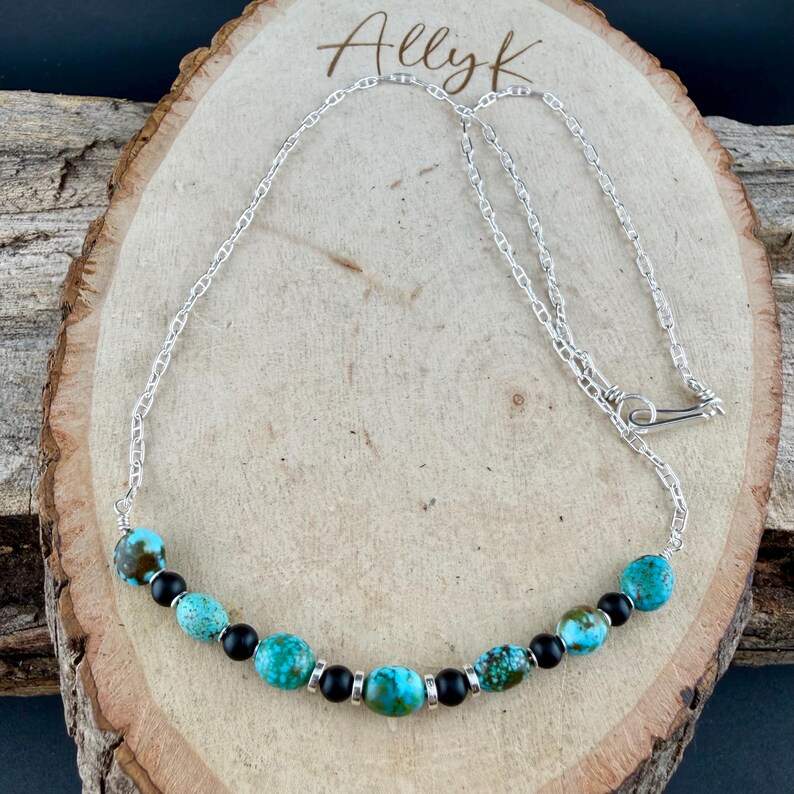 May include: A silver chain necklace with turquoise and black beads. The necklace features a series of round turquoise beads interspersed with smaller black beads. The chain is secured with a simple clasp. The name "Allyk" is written in cursive at the top.