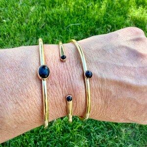 May include: A collection of gold-colored bracelets and rings. The bracelets have a simple, open design with black oval and round stones. The rings have a similar design, with a black stone at the end. The jewelry is displayed on a person's arm.