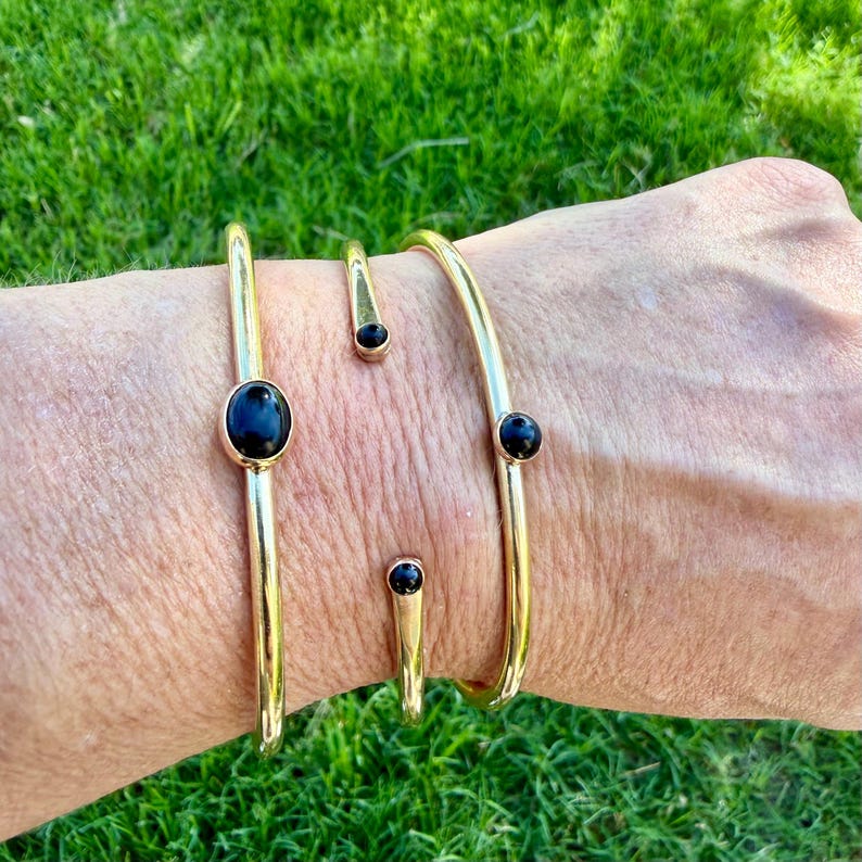 May include: A collection of gold-colored bracelets and rings. The bracelets have a simple, open design with black oval and round stones. The rings have a similar design, with a black stone at the end. The jewelry is displayed on a person's arm.
