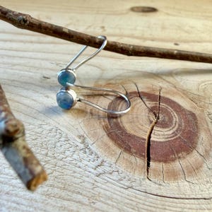 May include: A pair of silver earrings featuring round, iridescent blue cabochon stones. The earrings have a curved, hook-style wire design that loops through the stone setting, creating a minimalist and sleek appearance for the jewelry.