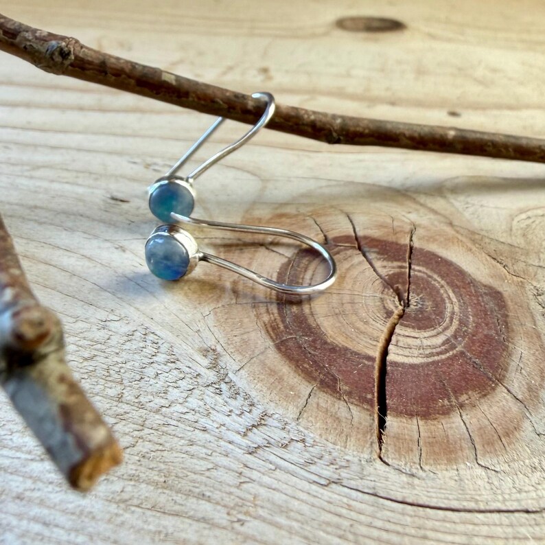 May include: A pair of silver earrings featuring round, iridescent blue cabochon stones. The earrings have a curved, hook-style wire design that loops through the stone setting, creating a minimalist and sleek appearance for the jewelry.