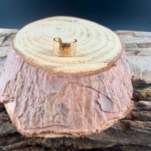May include: A gold-colored ring with an engraved floral pattern. The ring is a wide band style, and it is displayed on a wooden surface. The ring's design features swirling lines and curves, adding a touch of elegance to the jewelry.