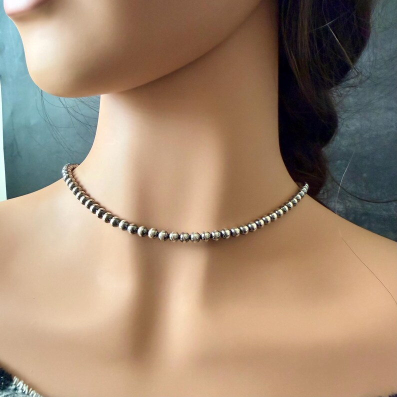 Pu&ograve; includere: Una collana girocollo con perline d'argento. La collana presenta piccole perline rotonde infilate insieme per formare una delicata catena. Le perline hanno un aspetto lucido e metallico. La collana &egrave; indossata intorno al collo.