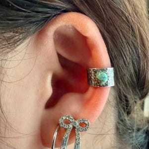 May include: A silver ear cuff with a textured design and a turquoise stone. Below, a silver earring with a bow design and clear stones. The jewelry is worn on an ear.