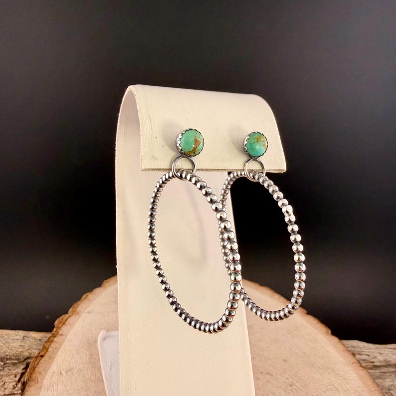 Puede incluir: Pendientes de aro plateados con piedras turquesas. Los pendientes presentan un dise&ntilde;o de cuentas alrededor de los aros y una peque&ntilde;a piedra turquesa en la parte superior. Se exhiben sobre un soporte blanco sobre un fondo oscuro.