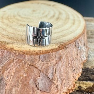 Puede incluir: Anillo de pu&ntilde;o plateado con superficie texturizada y dise&ntilde;o de cruz. El anillo est&aacute; abierto en la parte superior y se encuentra sobre una superficie de madera.