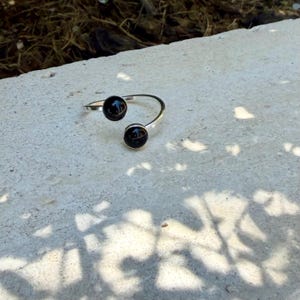 May include: An open-style ring featuring a thin, polished silver-tone metal band. The ring is accented with two smooth, round, black cabochon stones, one positioned at each end of the open band, creating a balanced and symmetrical appearance.