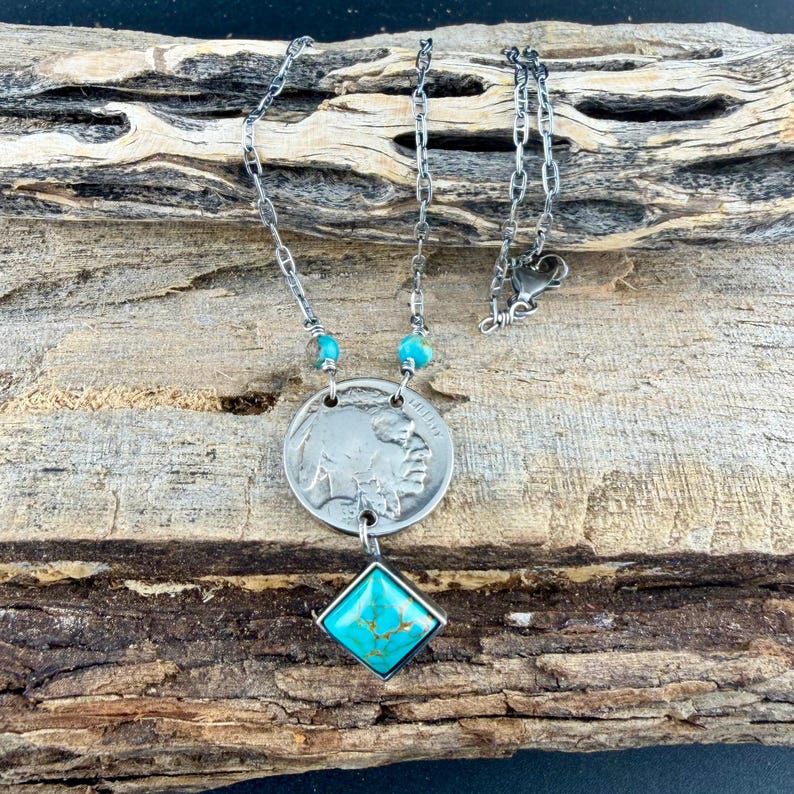 Op de afbeelding: Zilverkleurige ketting met een vintage munt-hanger met een buffel en een Indiaans profiel. De ketting bevat turquoise kralen en een vierkante turquoise stenen hanger. De ketting heeft een delicate schakelstijl en wordt gepresenteerd op een houten oppervlak.