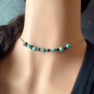 May include: A beaded choker necklace featuring turquoise and black beads strung on a silver-tone chain. The necklace is displayed on a person's neck, showcasing the jewelry's design and color contrast.