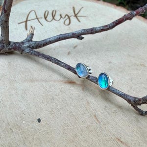May include: A pair of oval-shaped, iridescent gemstone stud earrings with silver settings. The earrings display shades of blue and green. The earrings are displayed on a wooden surface with a tree branch. The text "Allyk" is visible.