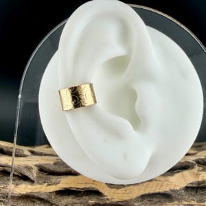 May include: A gold-colored ear cuff with an engraved floral pattern. The cuff is displayed on a white ear model, showcasing its design and fit. The jewelry piece is a stylish accessory.