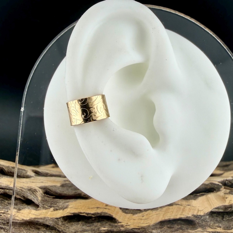 May include: A gold-colored ear cuff with an engraved floral pattern. The cuff is displayed on a white ear model, showcasing its design and fit. The jewelry piece is a stylish accessory.