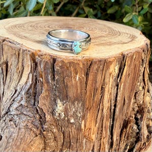 Puede incluir: Un anillo de banda de plata con acabado pulido y una piedra turquesa de forma ovalada con patrones de matriz oscuros. La banda de metal est&aacute; decorada con motivos grabados de flores u hojas que rodean el engaste central de la piedra.