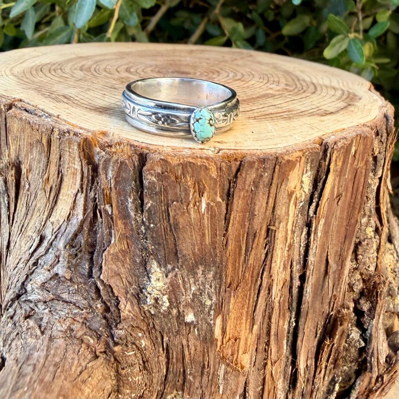 Puede incluir: Un anillo de banda de plata con acabado pulido y una piedra turquesa de forma ovalada con patrones de matriz oscuros. La banda de metal est&aacute; decorada con motivos grabados de flores u hojas que rodean el engaste central de la piedra.