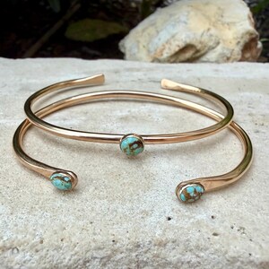 May include: Two gold-colored, open-ended cuff bracelets. Each bracelet features a small, round turquoise stone. The bracelets are displayed on a light-colored, textured surface, with a blurred natural background.