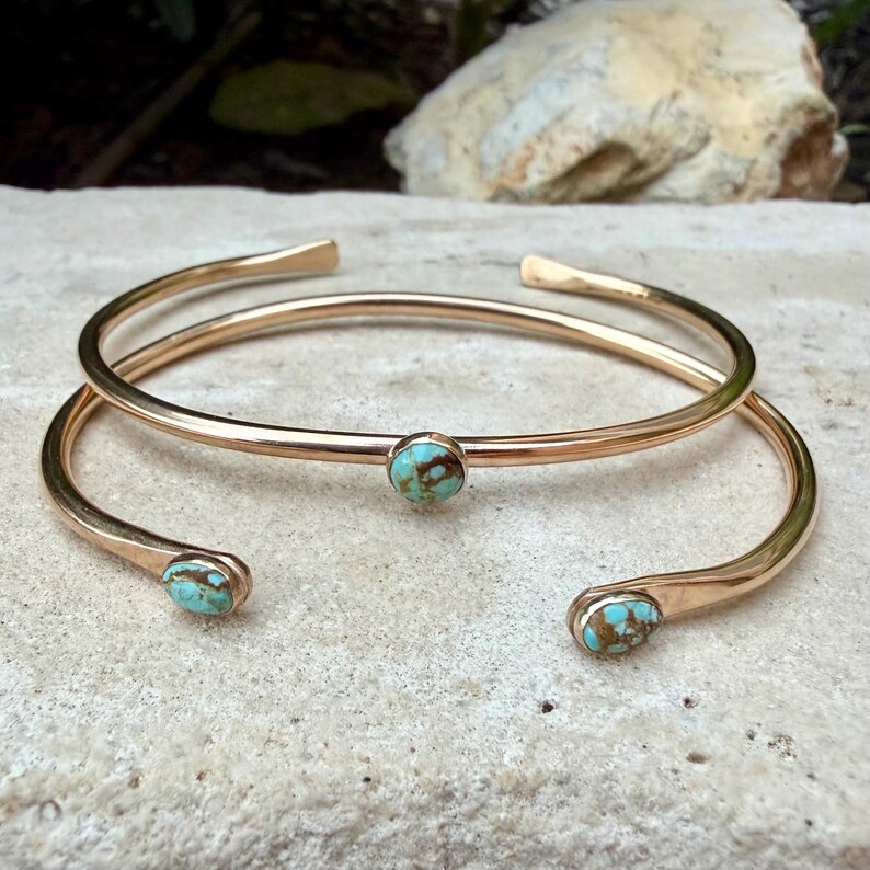 May include: Two gold-colored, open-ended cuff bracelets. Each bracelet features a small, round turquoise stone. The bracelets are displayed on a light-colored, textured surface, with a blurred natural background.