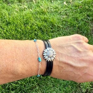 May include: A silver sunflower bracelet with a black leather band and a turquoise beaded bracelet. The sunflower bracelet has a detailed, textured design. The turquoise bracelet is made of small, round, blue-green beads. Both bracelets are worn on a wrist.