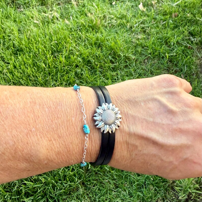May include: A silver sunflower bracelet with a black leather band and a turquoise beaded bracelet. The sunflower bracelet has a detailed, textured design. The turquoise bracelet is made of small, round, blue-green beads. Both bracelets are worn on a wrist.