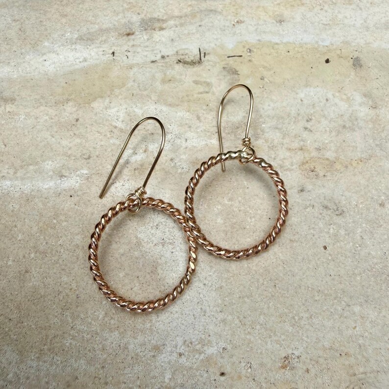 May include: A pair of rose gold-toned earrings featuring a circular hoop design with a twisted, rope-like texture. Each hoop is suspended from a simple, curved ear wire hook, creating a dangling silhouette with a metallic finish.