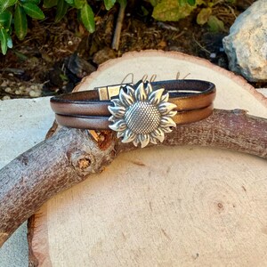 May include: Brown leather bracelet with a silver sunflower clasp. The bracelet features two bands of brown leather and a magnetic clasp. The sunflower clasp is detailed with textured petals and a central seed pattern.