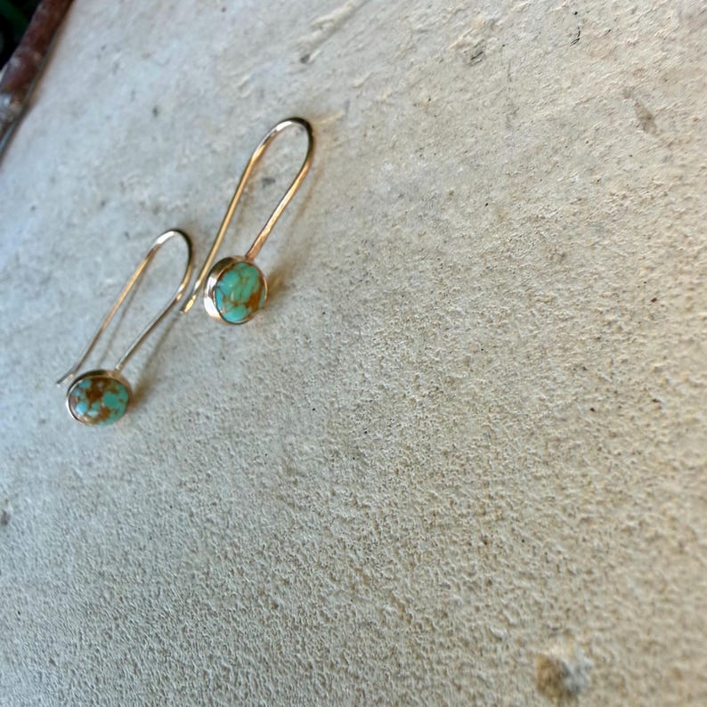 May include: A pair of drop earrings featuring round, light blue and brown marbled stones set in polished gold-tone metal. Each earring has a long, curved wire hook that extends upward from the circular stone setting, creating a simple, elegant profile.