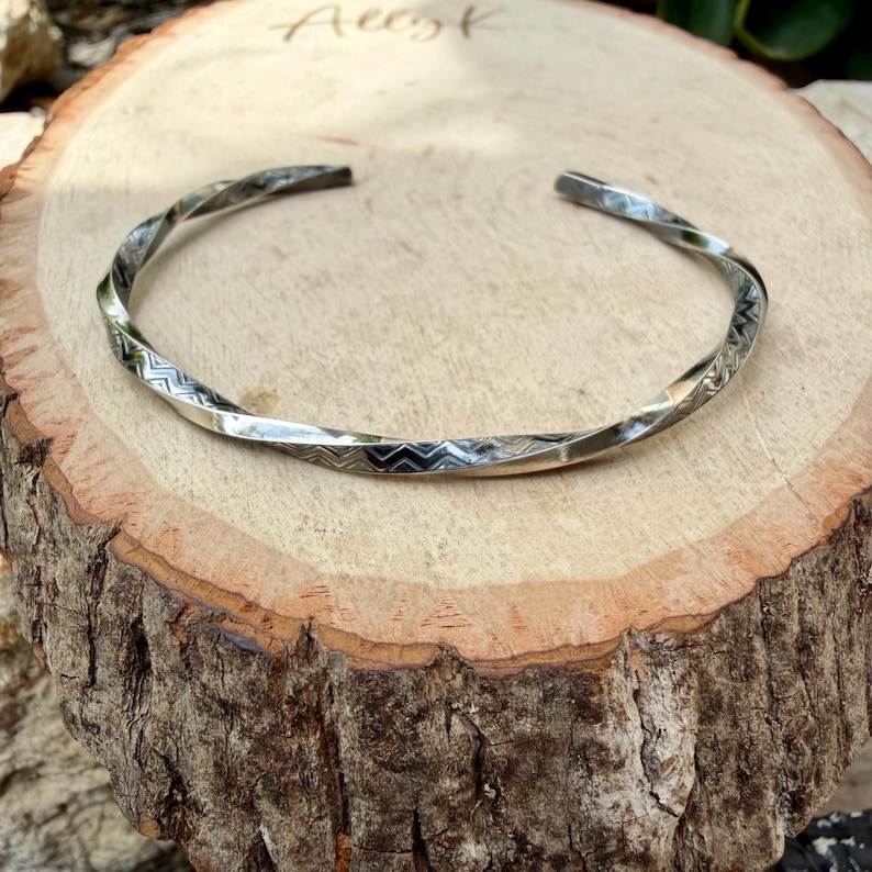 May include: A silver cuff bracelet with a twisted design and geometric patterns. The open-ended bracelet is displayed on a wooden surface, highlighting its texture and craftsmanship. The word "Ausk" is written on the wood.