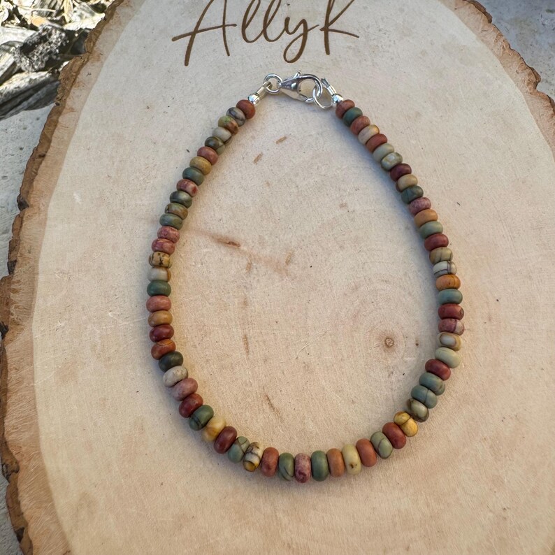 May include: A beaded bracelet with a variety of earth-toned beads, including shades of green, brown, yellow, and red. The bracelet is strung on a cord and secured with a silver clasp. The name "Allyk" is written in cursive at the top.