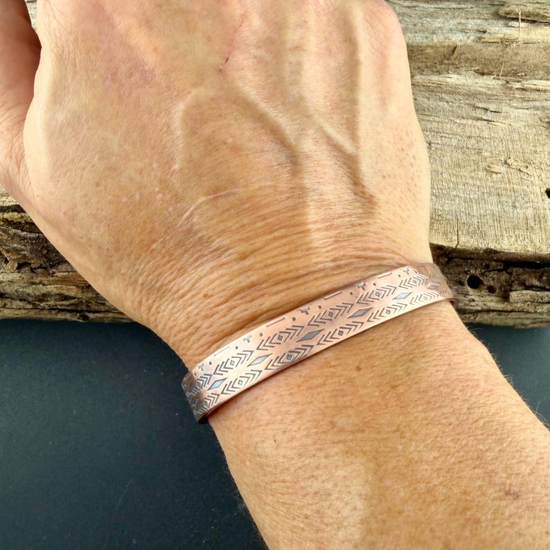 May include: A copper-toned metal cuff bracelet featuring a repeating engraved pattern of small diamonds, chevrons, and horizontal lines. The band has a smooth, polished finish and a consistent width, wrapping around the wrist with a simple open design.