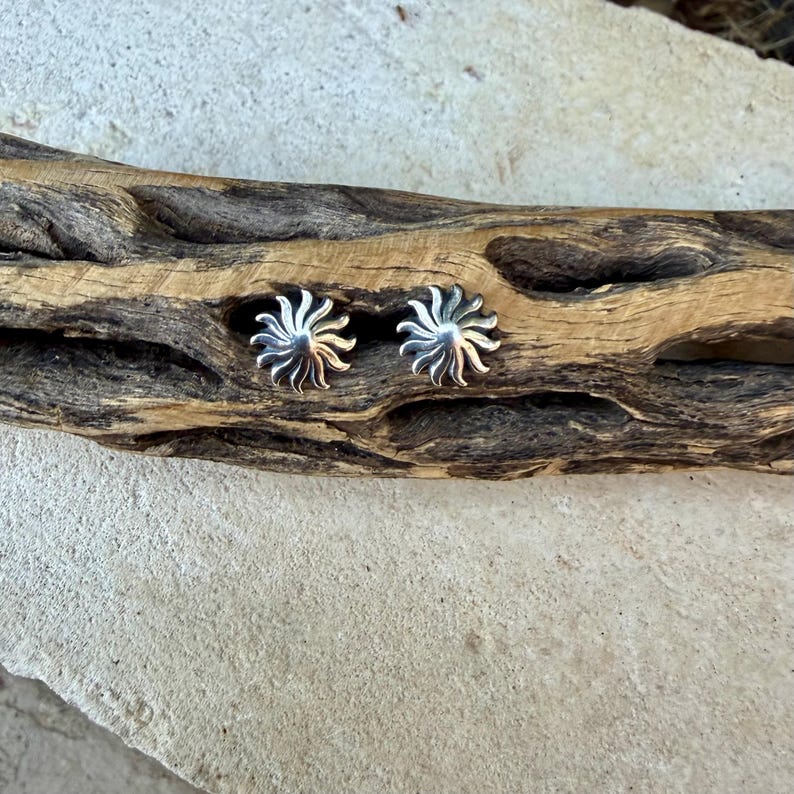 May include: A pair of silver stud earrings shaped like suns with radiating curved rays. Each earring features a polished metallic finish with a central dome and multiple slender, pointed extensions that fan out in a circular pattern.