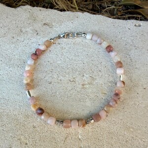May include: A beaded bracelet featuring small, faceted, light pink and tan stone beads arranged in a circular pattern. The bracelet includes several small, polished silver-toned metal spacer beads and a silver-toned lobster claw clasp closure.