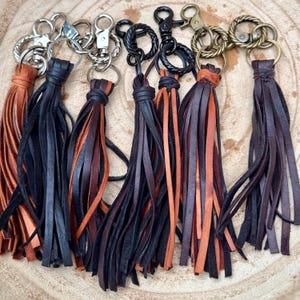 May include: A collection of leather keychains with various metal hardware. The keychains feature long, fringed tassels in shades of brown, black, and orange. The hardware includes silver, black, and brass-toned clasps and rings. The keychains are arranged on a wooden surface.