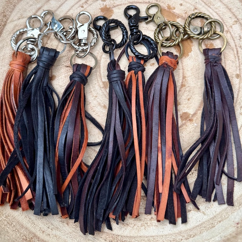 May include: A collection of leather keychains with various metal hardware. The keychains feature long, fringed tassels in shades of brown, black, and orange. The hardware includes silver, black, and brass-toned clasps and rings. The keychains are arranged on a wooden surface.