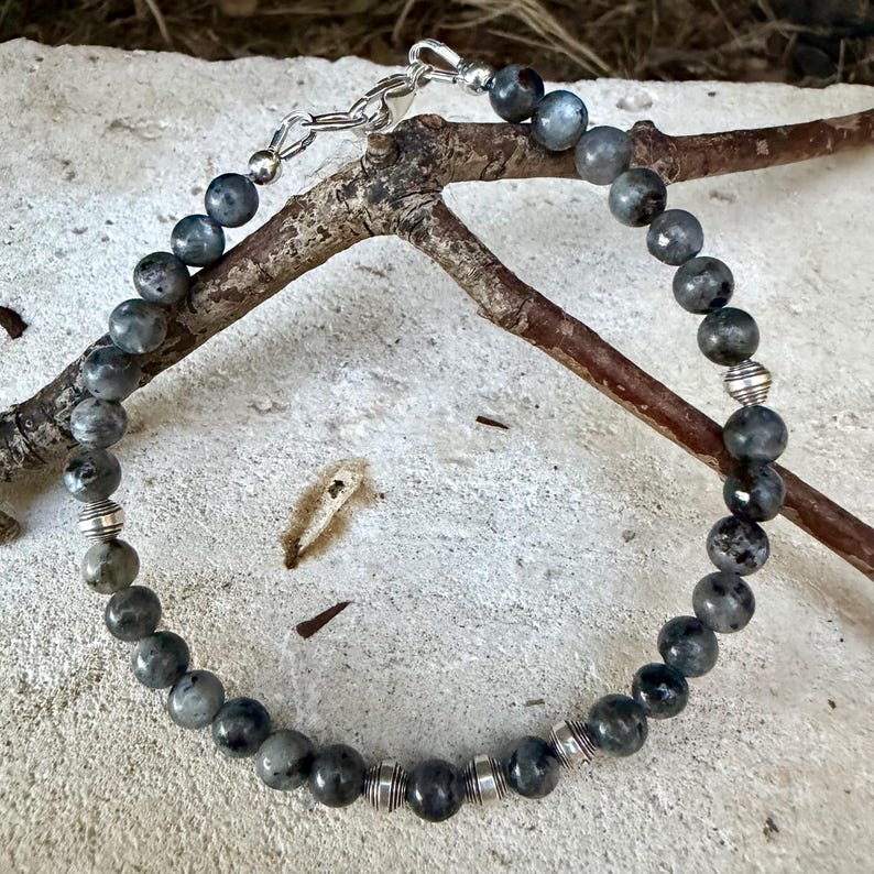 May include: A beaded bracelet featuring round, dark grey labradorite stones interspersed with small, ribbed silver-toned metal spacers. The bracelet is secured with a silver-toned lobster claw clasp and a short extender chain.