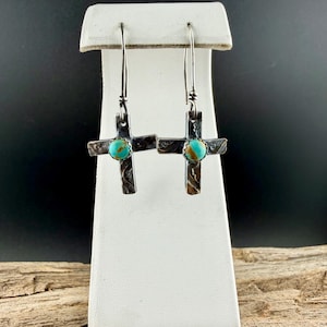 May include: Silver-tone cross earrings with turquoise-colored stone accents. The crosses have a textured, etched design and dangle from long, thin hooks. The earrings are displayed on a white stand against a dark background.