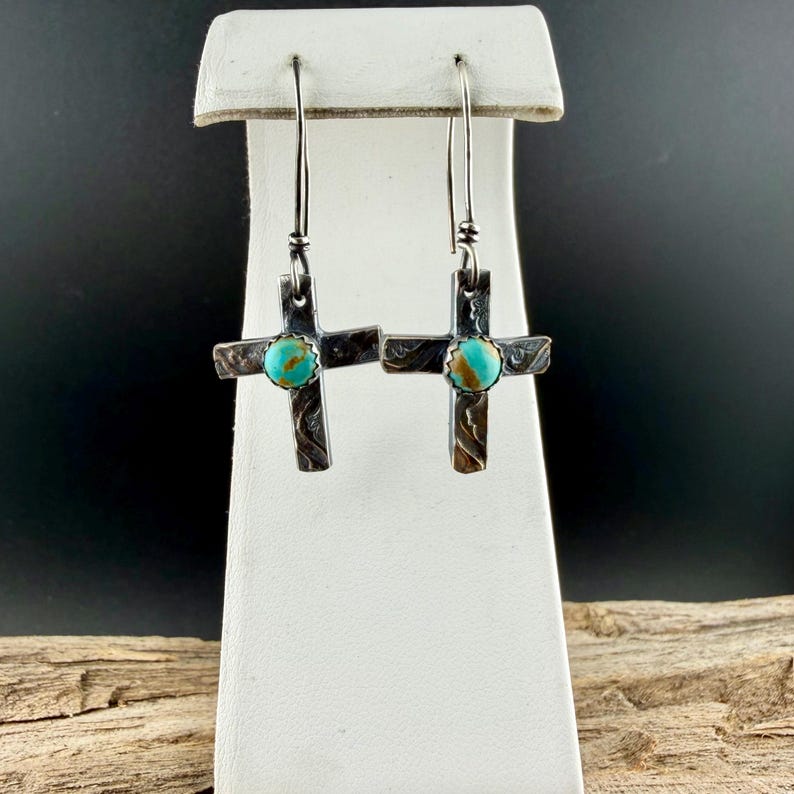 May include: Silver-tone cross earrings with turquoise-colored stone accents. The crosses have a textured, etched design and dangle from long, thin hooks. The earrings are displayed on a white stand against a dark background.