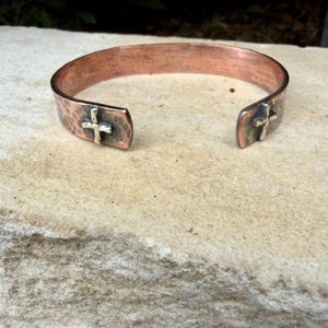 May include: A copper cuff bracelet featuring a hammered, textured surface and an open design. Each end of the bracelet is adorned with a small, silver-toned metal cross emblem, providing a distinct contrast to the warm copper finish of the band.