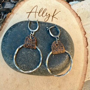 May include: A pair of silver hoop earrings with a twisted design. Each earring features a brown leather accent and a hinged clasp. The earrings are displayed on a dark gray stone, with the text "Allyk" in the background.
