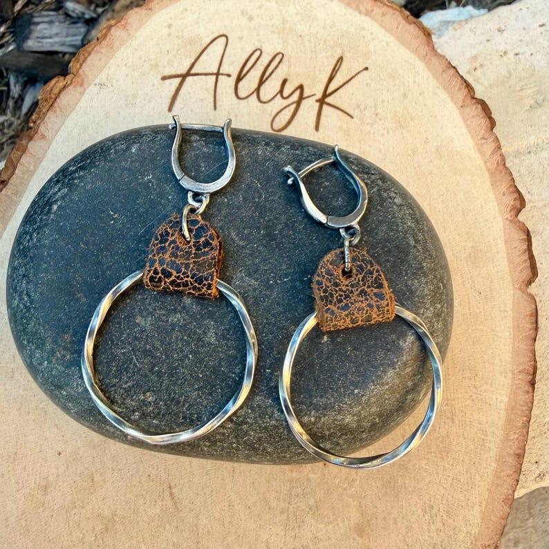 May include: A pair of silver hoop earrings with a twisted design. Each earring features a brown leather accent and a hinged clasp. The earrings are displayed on a dark gray stone, with the text "Allyk" in the background.