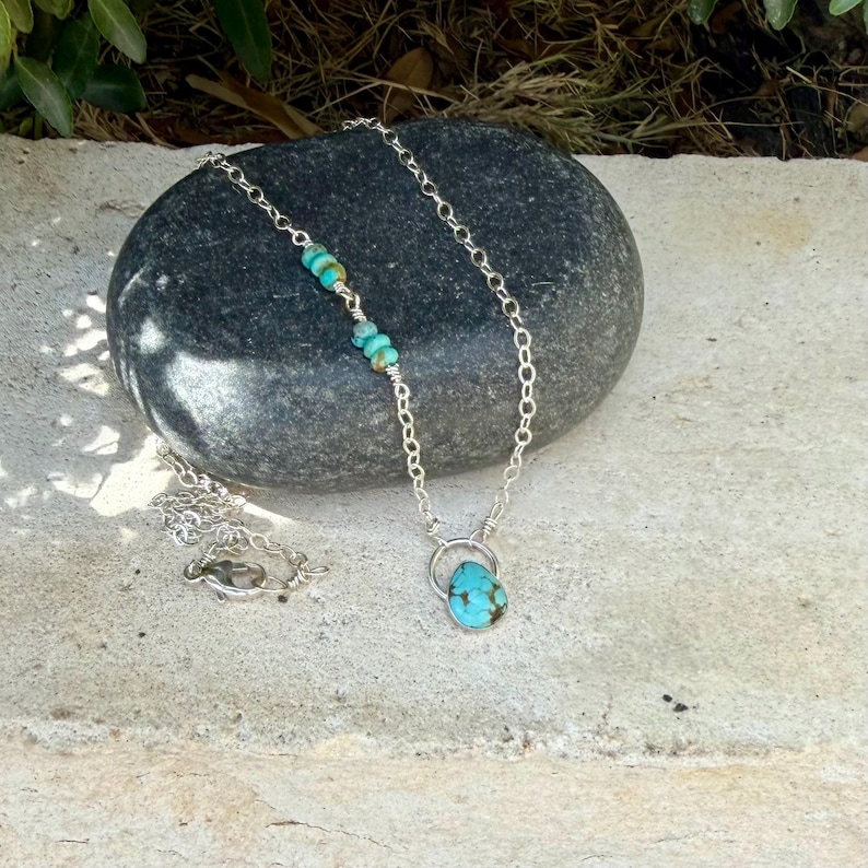 May include: A silver chain necklace featuring a teardrop-shaped turquoise pendant and small turquoise beads integrated into the chain. The necklace includes a lobster claw clasp and is displayed on a dark gray stone surface with a light stone base.