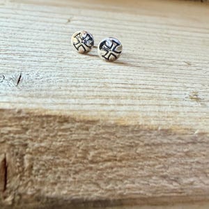 May include: A pair of small, round silver stud earrings. Each earring features a polished metallic surface with a dark, recessed cross symbol engraved in the center, creating a distinct contrast against the bright, reflective silver finish.
