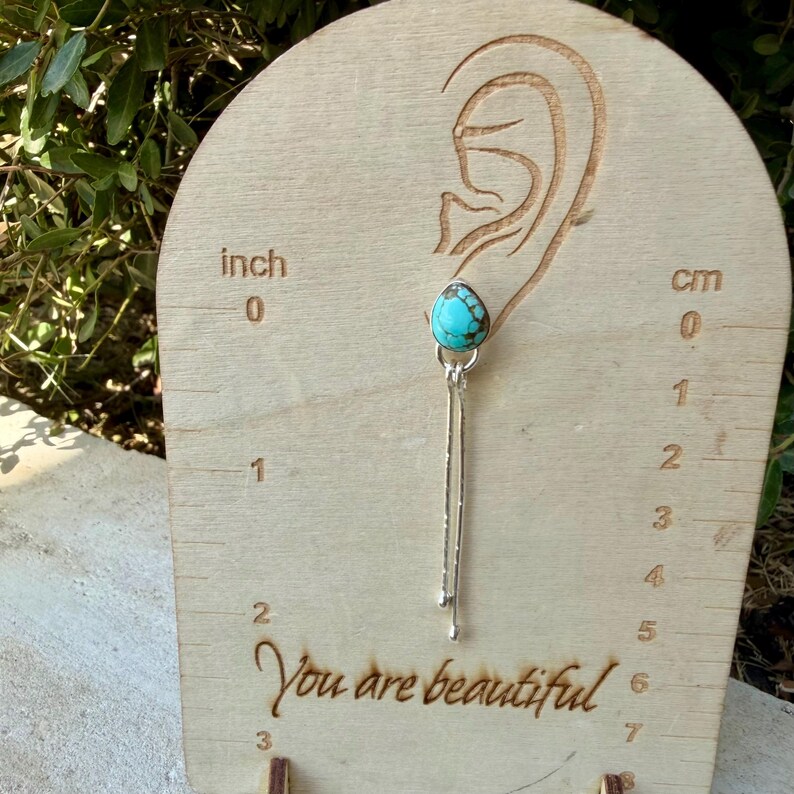 May include: A silver earring featuring a teardrop-shaped turquoise stone with dark matrix patterns, suspended from a silver post with two thin, dangling silver chains. The earring is displayed on a wooden card with inch and cm markings.