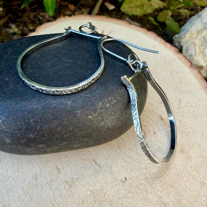 May include: A pair of silver teardrop earrings with a textured, etched design. The earrings are displayed on a dark gray stone, showcasing their intricate details and craftsmanship. The earrings have a hook closure.