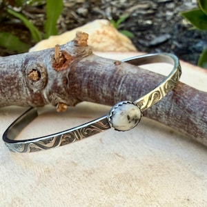 May include: A silver bangle bracelet with an engraved swirl pattern. The bracelet features a round, white stone with black speckles set in a scalloped bezel. The bracelet is displayed on a wooden branch.