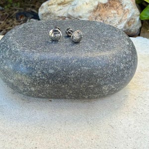 May include: A pair of small, round, silver-toned stud earrings featuring a central diamond-shaped engraving surrounded by fine, etched line details. The earrings have a metallic finish and are designed with a post-back closure mechanism.