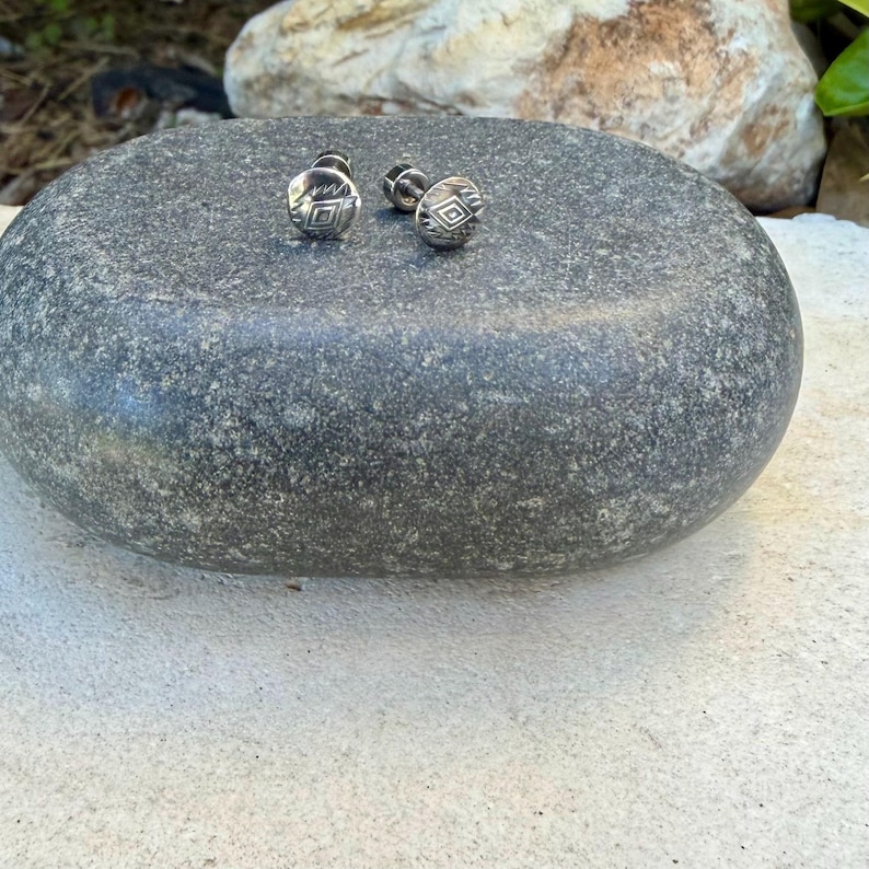 May include: A pair of small, round, silver-toned stud earrings featuring a central diamond-shaped engraving surrounded by fine, etched line details. The earrings have a metallic finish and are designed with a post-back closure mechanism.