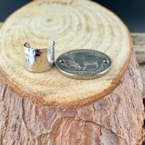 May include: A silver-colored ear cuff with a hammered texture and a vintage five-cent coin featuring a buffalo. The coin is oval-shaped and has the words "FIVE CENTS" engraved on it. The items are displayed on a wooden surface.